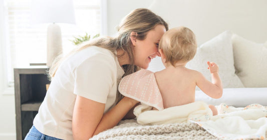 Mom gently cleaning baby’s back on bed with a Cami baby washcloth, capturing a soft and heartfelt bonding moment.