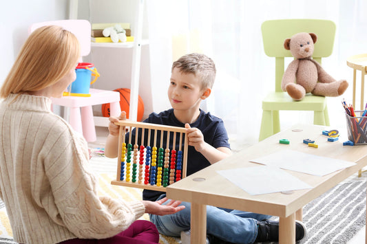A woman and a young boy sitting at a table playing with colorful wooden blocks in a bright, cozy room with toys in the background.